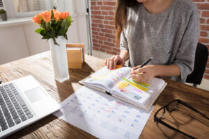 Foto de uma mulher em uma mesa, com vaso de flor, notbook e fazendo planejamento.